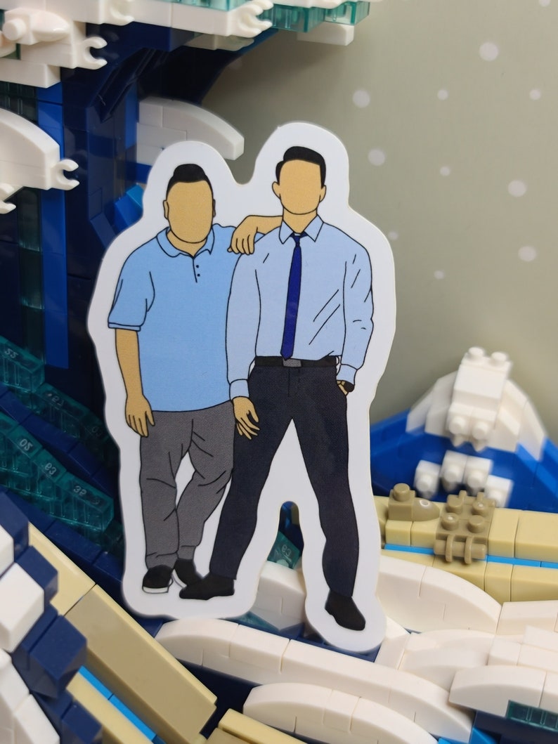May include: A sticker featuring two figures in casual attire. One figure wears a light blue polo shirt and gray pants, while the other is in a light blue button-down shirt, dark pants, and a blue tie. The sticker has a white border.