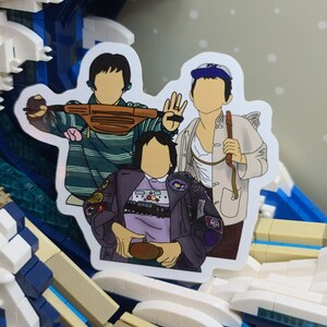 May include: A sticker featuring a cartoon illustration of three people. One person is wearing a striped shirt and holding a brown fanny pack. Another is wearing a white jacket and holding a stick. The third person is wearing a purple jacket.