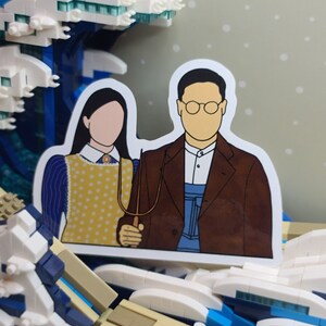 May include: A sticker featuring a parody of the American Gothic painting, with the figures' faces blank. The figures are holding a pitchfork. The sticker is set against a backdrop of a Lego wave.