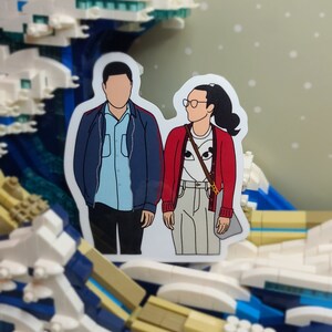 May include: A sticker featuring a couple, with faceless illustrations, standing side-by-side. The woman wears a red cardigan, glasses, and a ponytail. The man wears a blue jacket. The sticker is set against a Lego wave background.