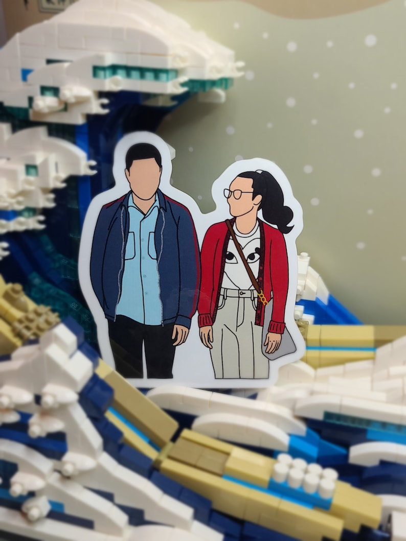 May include: A sticker featuring a couple, with faceless illustrations, standing side-by-side. The woman wears a red cardigan, glasses, and a ponytail. The man wears a blue jacket. The sticker is set against a Lego wave background.