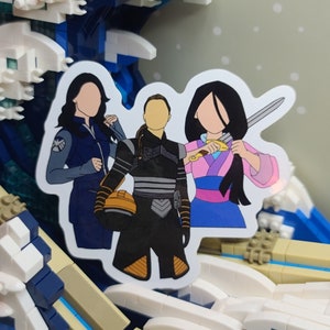 May include: A sticker featuring three stylized female figures. One wears a blue jacket with a shield emblem, another a black and gray outfit with an orange helmet, and the third a pink and blue kimono holding a sword. The sticker is set against a wave-like background.