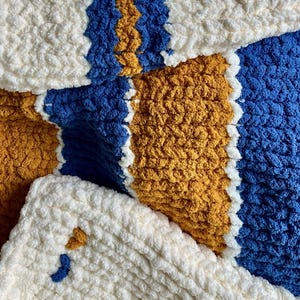 Pode incluir: Close-up de um cobertor macio e texturizado. O cobertor apresenta um padrão de listras brancas, azuis e douradas. O fio grosso cria uma aparência aconchegante e convidativa. O esquema de cores é brilhante e alegre.