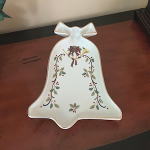 May include: A white ceramic serving dish shaped like a bell with a gold-trimmed edge. The center features a decorative illustration of a gold horn tied with a red ribbon, surrounded by green holly leaves and red berries trailing down the sides.