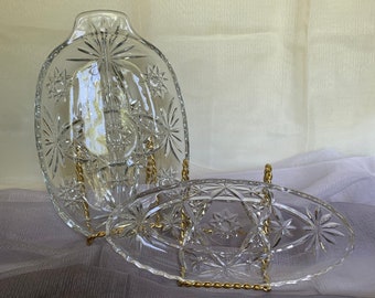 May include: Two clear glass serving dishes with a starburst pattern. The dishes are oval shaped and have a raised edge. They are displayed on a gold metal stand.