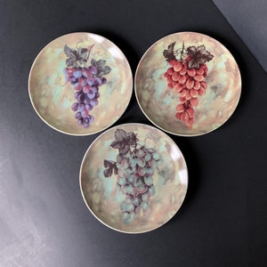 May include: Three decorative ceramic plates with a watercolor design of grapes on a light yellow background. The grapes are purple, red, and green.