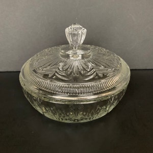 May include: A clear glass candy dish with a matching lid. The dish features a pressed glass pattern with vertical fluting on the base and a decorative starburst design on the lid. A small, faceted glass knob sits atop the center of the lid.