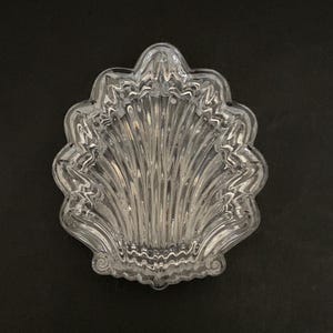 May include: A clear, glass seashell-shaped dish with a scalloped edge and vertical ridged detailing. The dish is set against a black background, highlighting its transparent nature and intricate design. A decorative piece for serving or display.