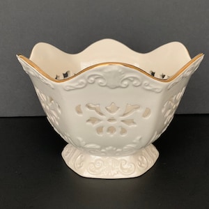 May include: Cream-colored ceramic bowl with a scalloped rim and a gold trim. The bowl features intricate cut-out designs and rests on a decorative pedestal base. A classic piece for serving or display.