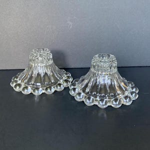 May include: Two clear glass candle holders. Each has a flared, ribbed body and a circular base adorned with small, round glass beads. The top has a small opening for a candle. The candle holders are displayed on a dark surface.