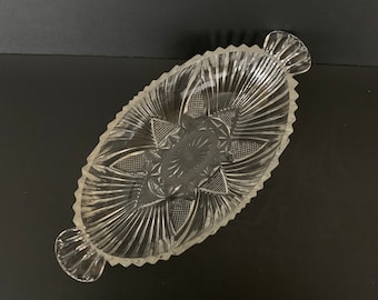 May include: A clear glass serving dish with a decorative pattern and two handles. The dish is oval shaped and has a raised center.