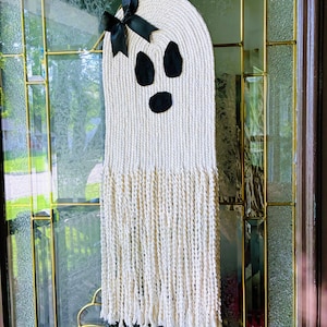 May include: A handmade, cream-coloured ghost decoration with a black bow and black facial features. The ghost is made of woven material with a fringed bottom. It is hanging on a glass door.
