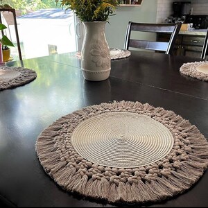 以下が含まれることがあります： 濃い木製のテーブルの上に、フリンジのトリムが付いた丸いベージュのプレースマットが3つあります。プレースマットは織り素材で作られており、微妙な質感があります。