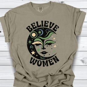 Może przedstawiać: Szałwiowa zielona koszulka z napisem "BELIEVE WOMEN" w kolorze czarnym, otaczającym wzór półksiężyca. Grafika księżyca zawiera gwiazdy, twarz oraz zielone i niebieskie akcenty. Koszulka jest wykonana z miękkiego materiału.