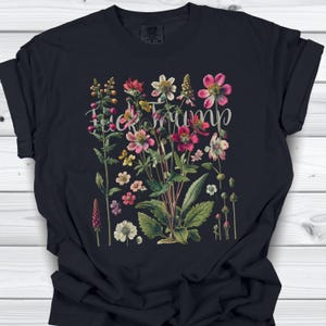 May include: Black t-shirt featuring a floral design with the text "Fuck Trump" in a cursive font. The flowers are in shades of pink, red, yellow, and white, with green leaves. The shirt is made of a soft material.