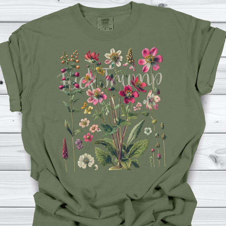 May include: Olive green t-shirt featuring a floral design with pink, red, and white flowers. The text "F*ck Trump" is printed in a cursive font across the flowers. The shirt is made of a soft, comfortable material.