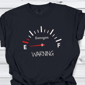 May include: A black t-shirt featuring a white and red graphic of a fuel gauge. The gauge displays the word Estrogen, with a red needle pointing toward the E. Below the gauge, the word WARNING is printed in white capital letters.