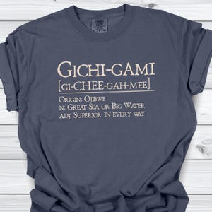 Peut inclure: Un t-shirt bleu ardoise avec un texte couleur crème qui indique GICHI-GAMI [GI-CHEE-GAH-MEE] ORIGIN: OJIBWE N: GREAT SEA OR BIG WATER ADJ: SUPERIOR IN EVERY WAY. Le t-shirt présente une encolure ronde et des manches courtes avec une étiquette visible.
