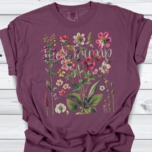 May include: A maroon t-shirt featuring a floral design with the text "F*ck Trump" in a cursive font. The floral arrangement includes various flowers in shades of pink, red, yellow, and white, with green leaves.