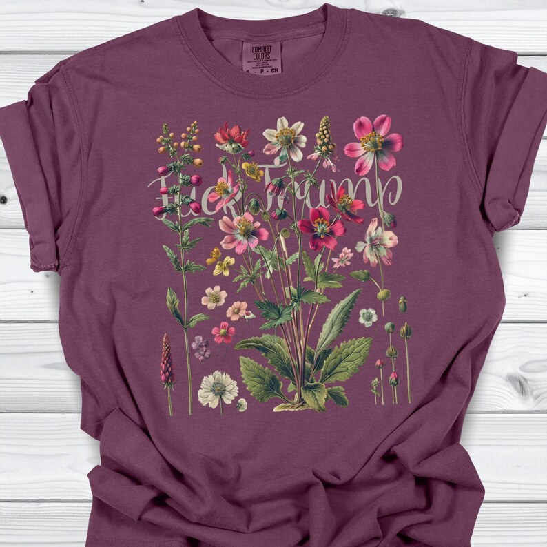 May include: A maroon t-shirt featuring a floral design with the text "F*ck Trump" in a cursive font. The floral arrangement includes various flowers in shades of pink, red, yellow, and white, with green leaves.