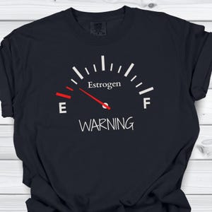 May include: Black t-shirt with a white gauge graphic that reads "Estrogen" and "Warning." The gauge needle is red and points to the "E" side. The shirt is made of a soft material.