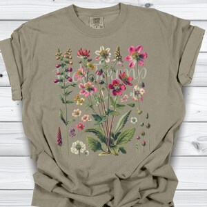 May include: A tan t-shirt featuring a botanical illustration of various flowers in shades of pink, red, white, and yellow. The floral design includes detailed leaves and stems. The text "Bloom" is written across the flowers.