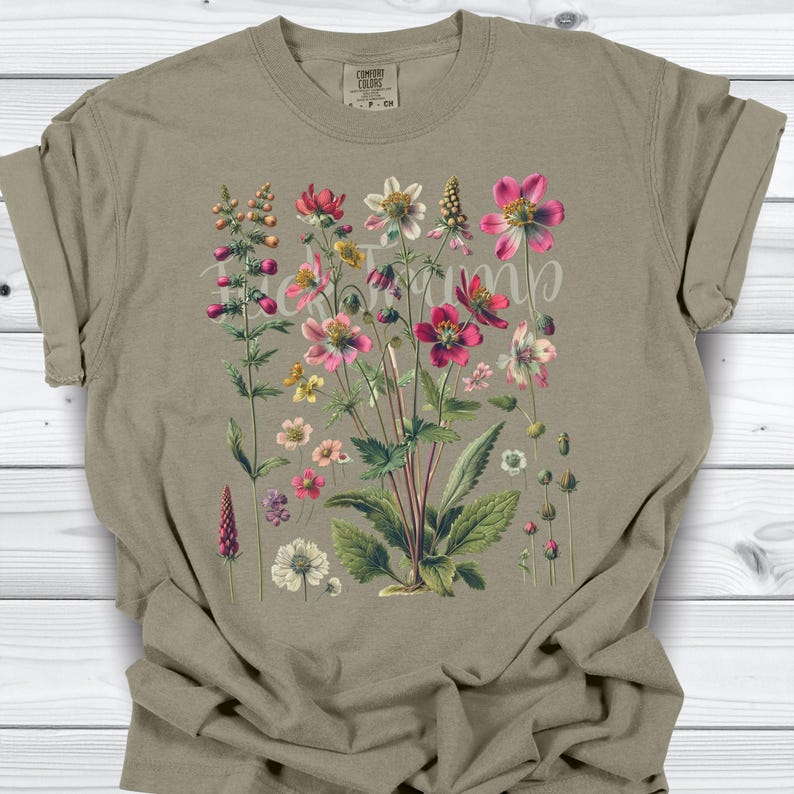 May include: A tan t-shirt featuring a botanical illustration of various flowers in shades of pink, red, white, and yellow. The floral design includes detailed leaves and stems. The text "Bloom" is written across the flowers.