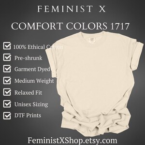 May include: Cream-colored t-shirt with rolled sleeves. The shirt features the text "FEMINIST X" and "COMFORT COLORS 1717". Additional text includes "100% Ethical Cotton", "Pre-shrunk", "Garment Dyed", "Medium Weight", "Relaxed Fit", "Unisex Sizing", and "DTF Prints".