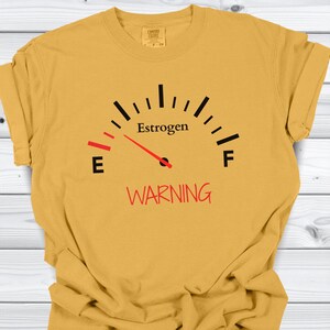 May include: Mustard yellow t-shirt with a gauge graphic. The gauge reads "Estrogen" with a red needle pointing towards "E" and the word "WARNING" in red below. The shirt has a Comfort Colors tag.