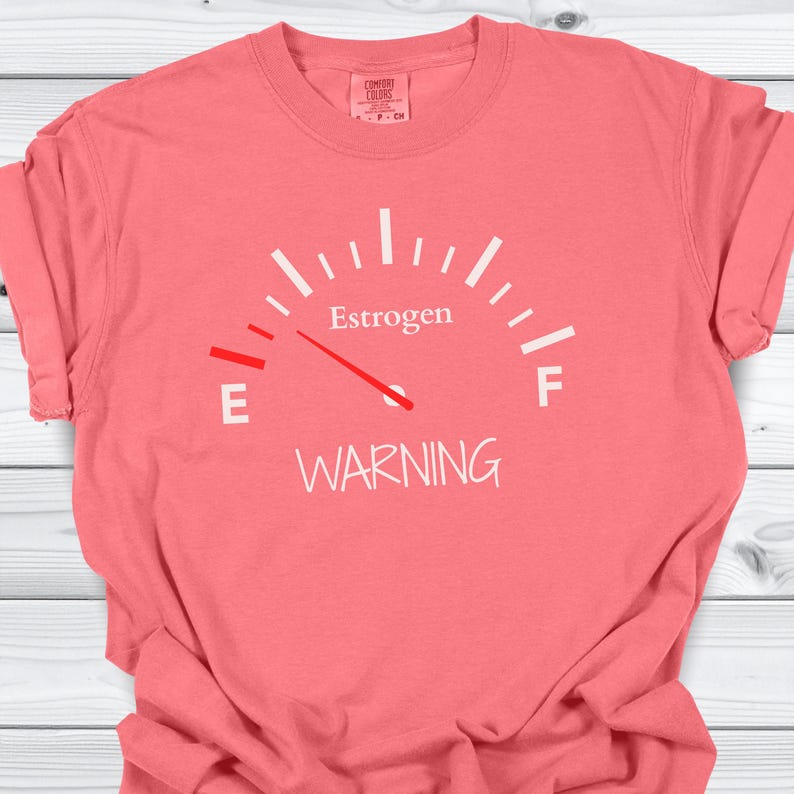 May include: Coral-colored t-shirt with a white graphic of a fuel gauge. The gauge reads "Estrogen" with the needle pointing towards "E" and the word "WARNING" below. The shirt is made of a soft material.