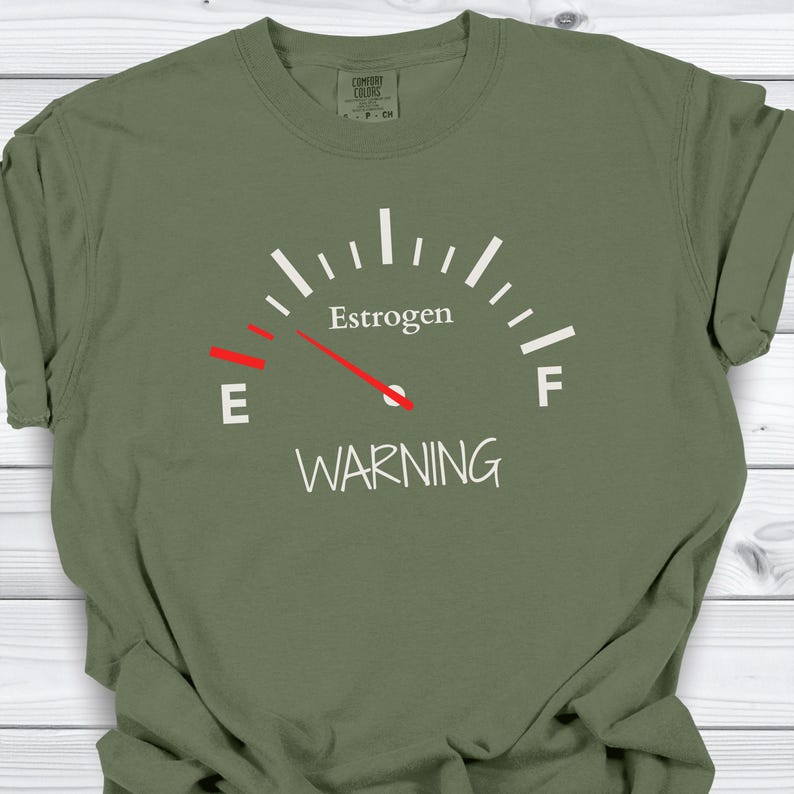 May include: Olive green t-shirt with a graphic of a gauge reading "Estrogen" with the needle pointing towards "E" and the word "WARNING" below. The shirt has a crew neck and short sleeves.