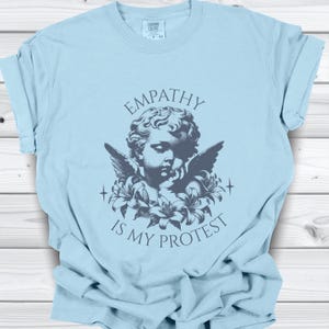 May include: Light blue t-shirt with the text "EMPATHY IS MY PROTEST" in a dark grey font. The graphic features a cherub with wings and flowers. The shirt is made of a soft material.