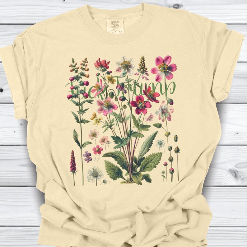 May include: Cream-colored t-shirt featuring a floral design with the word "Heck Yeah" in green script. The graphic includes various flowers in shades of pink, red, and white, with green leaves. The shirt is made of a soft material.