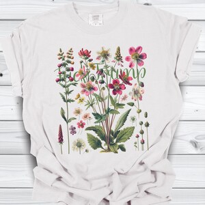 May include: White t-shirt featuring a botanical illustration of various flowers in shades of pink, red, and yellow, with green leaves and stems. The word "Heirloom" is written in green script across the design. The shirt is made of a soft material.
