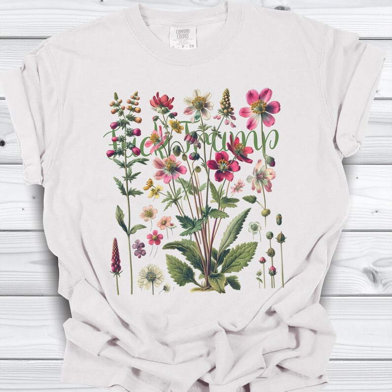 May include: White t-shirt featuring a botanical illustration of various flowers in shades of pink, red, and yellow, with green leaves and stems. The word "Heirloom" is written in green script across the design. The shirt is made of a soft material.