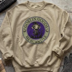 May include: A beige crewneck sweatshirt featuring a circular graphic on the chest. The design includes a purple center with a black illustration of a scarecrow-like figure, surrounded by a light green border with the text GOBLIN GOODS MERCANTILE.