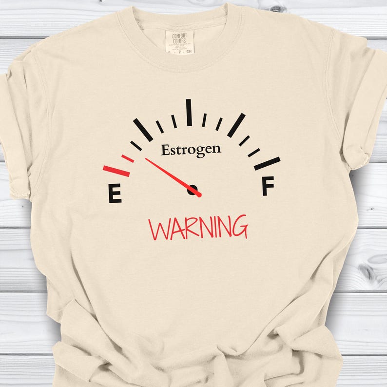 May include: Cream-colored t-shirt featuring a gauge design with the word "Estrogen" above a red needle pointing towards "E". Below the gauge, the word "WARNING" is printed in red. The shirt has a crew neck and short sleeves.