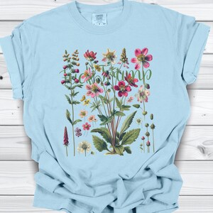May include: Light blue t-shirt featuring a botanical illustration of various flowers in shades of pink, red, yellow, and white, with green leaves. The word "Tramp" is written in a cursive font across the flowers.