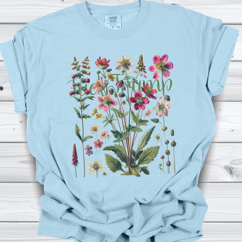 May include: Light blue t-shirt featuring a botanical illustration of various flowers in shades of pink, red, yellow, and white, with green leaves. The word "Tramp" is written in a cursive font across the flowers.