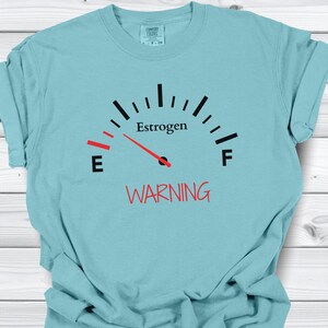 May include: A light blue t-shirt with a gauge graphic that reads "Estrogen" with a needle pointing to the left. The word "WARNING" is printed in red below the gauge.