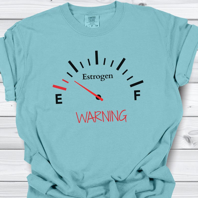 May include: A light blue t-shirt with a gauge graphic that reads "Estrogen" with a needle pointing to the left. The word "WARNING" is printed in red below the gauge.