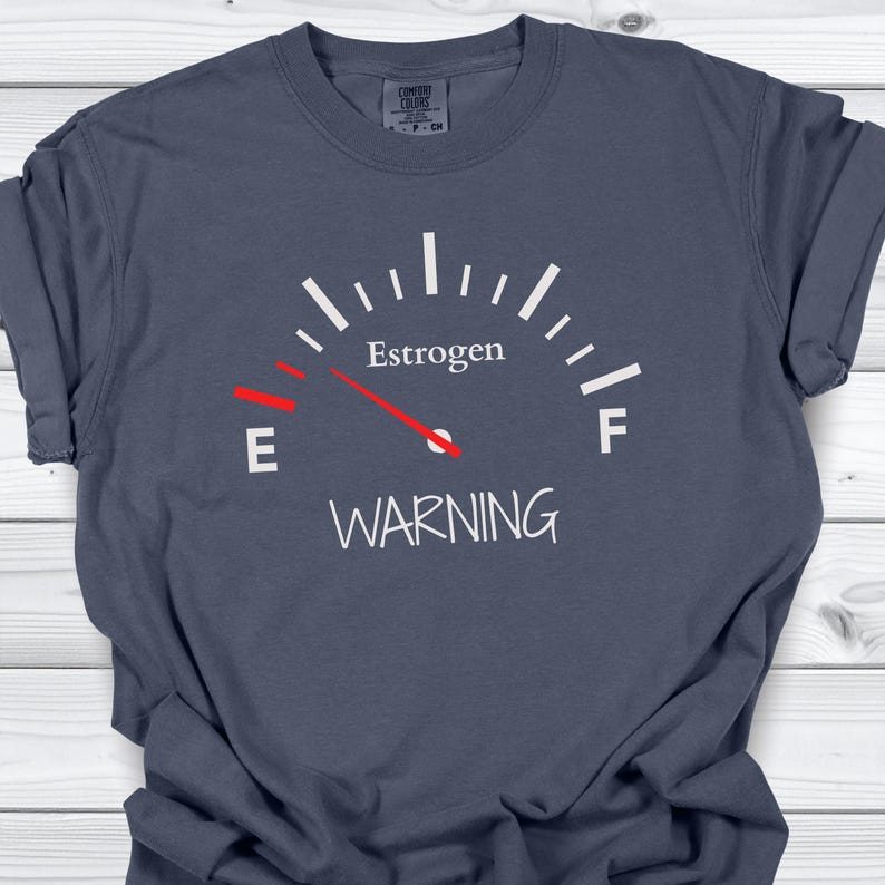 May include: A navy blue t-shirt with a gauge graphic that reads "Estrogen" with the needle pointing to "E" and the word "WARNING" below. The shirt is made of a soft material.