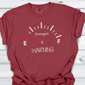 May include: A maroon t-shirt features a white gauge design with the word "Estrogen" at the top and "WARNING" at the bottom. The gauge needle points towards the "E" for empty.