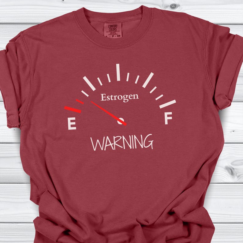 May include: A maroon t-shirt features a white gauge design with the word "Estrogen" at the top and "WARNING" at the bottom. The gauge needle points towards the "E" for empty.