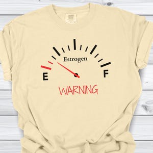 May include: Cream-colored t-shirt featuring a gauge design with the word "Estrogen" above a red needle pointing to "E". Below the gauge, the word "WARNING" is printed in red. The shirt is made of a soft, comfortable material.