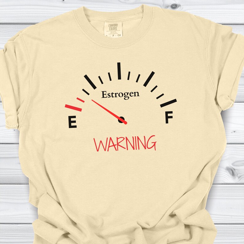 May include: Cream-colored t-shirt featuring a gauge design with the word "Estrogen" above a red needle pointing to "E". Below the gauge, the word "WARNING" is printed in red. The shirt is made of a soft, comfortable material.