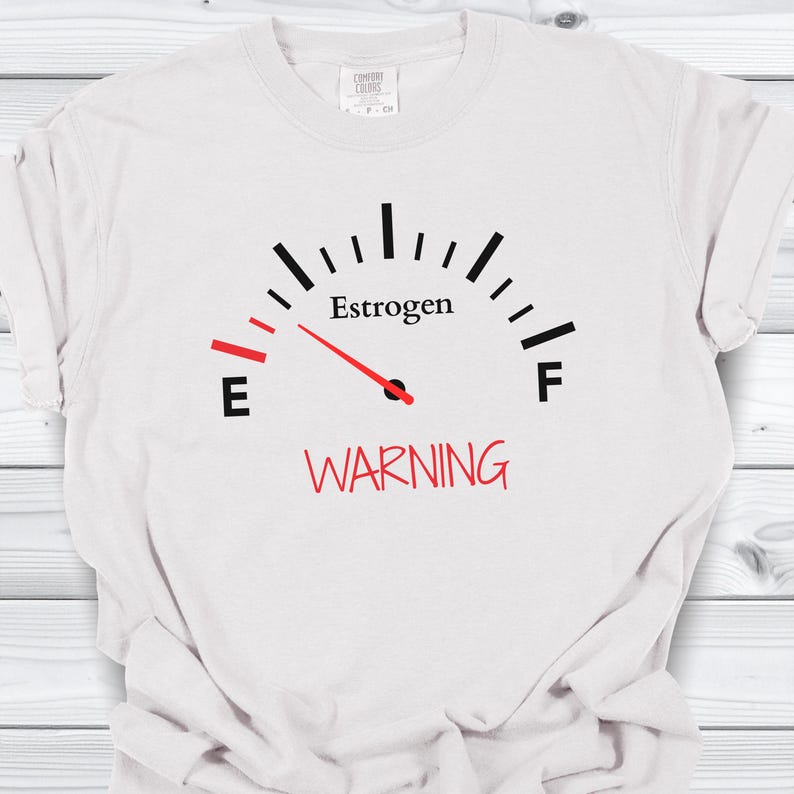 May include: White t-shirt with a black and red graphic. The graphic resembles a fuel gauge, with the word "Estrogen" above the gauge and "WARNING" below. The gauge needle points towards the "E".