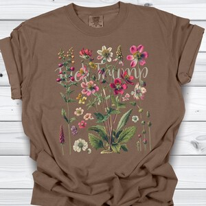 May include: Brown t-shirt featuring a floral design with the text "F*ck Trump" in a script font. The flowers are in shades of pink, yellow, and white, with green leaves. The shirt is a Comfort Colors brand.