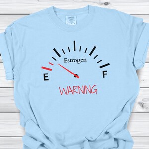 May include: Light blue t-shirt with a gauge graphic. The gauge reads "Estrogen" with a red needle pointing towards "E" and the word "WARNING" in red below the gauge.