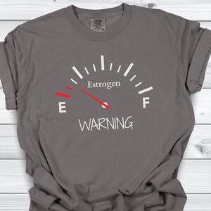 May include: Gray t-shirt with a white graphic of a gauge reading "Estrogen" with a red needle pointing towards "E" and the word "WARNING" below.
